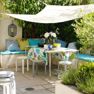 Outdoor terrace room with white fabric canopy, built in concrete seating, blue seat cushions, table and stools with plant print table cloth, patio