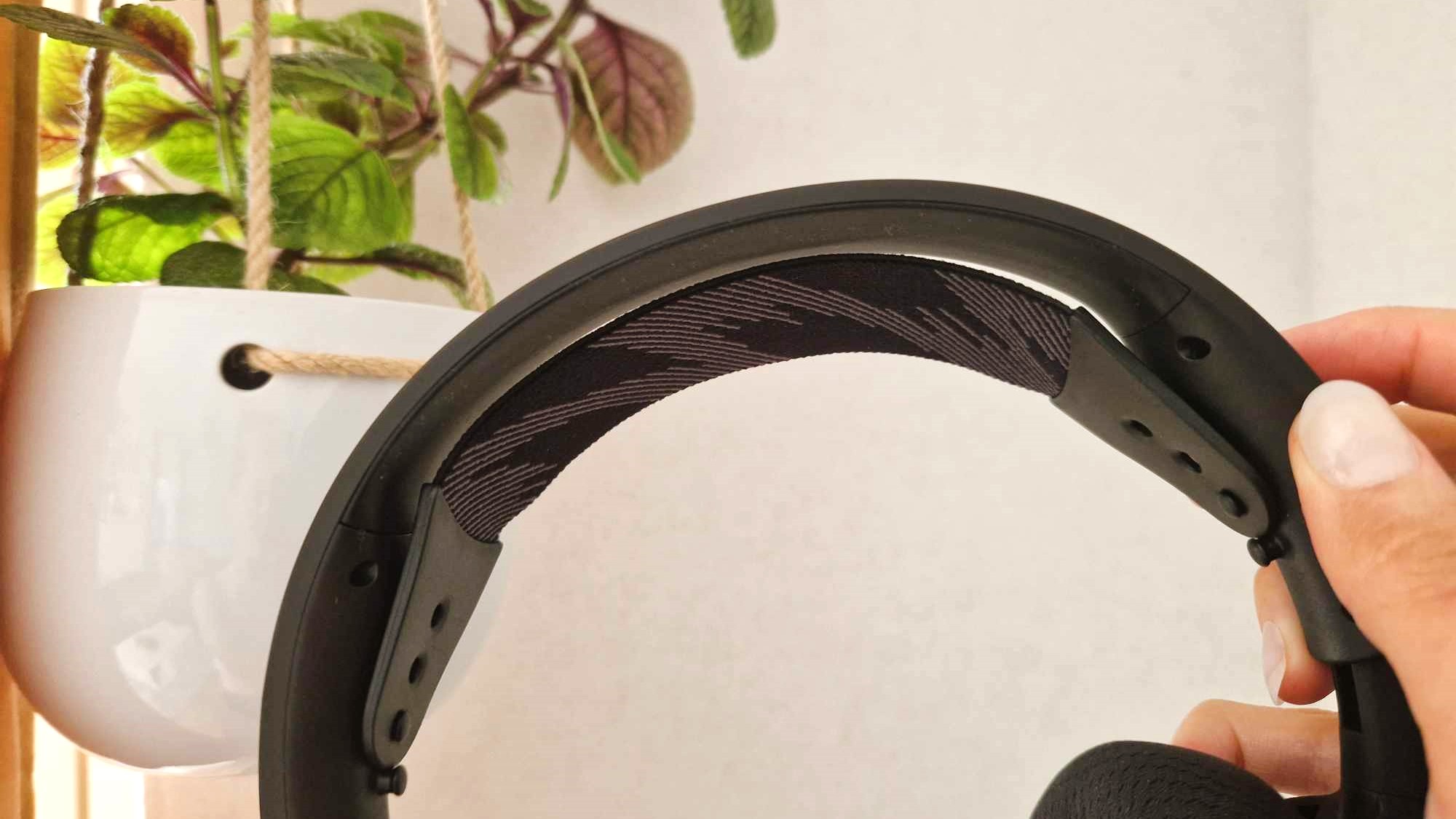 En hand håller upp ett par SteelSeries Arctis Nova 4 framför en hängande krukväxt vid en vit vägg, med fokus på huvudbandet.