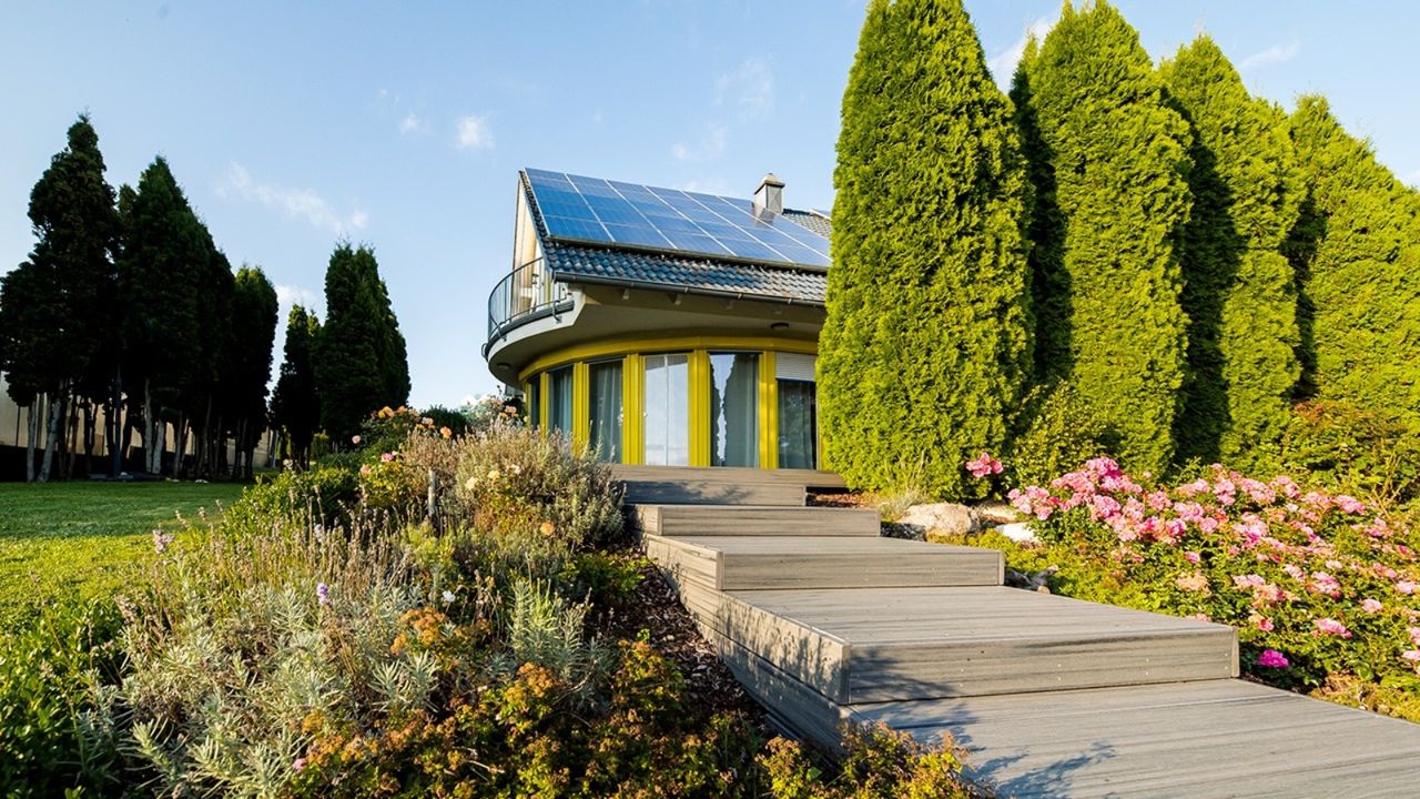 Bright photo of wooden stair pathway leading to large house with big windows and solar panels, surrounded by trees and bushes, with a bright blue sky above.