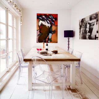 an all-white dining area in a garage conversion with floor to ceiling windows, artwork on the walls, and a wooden dining table