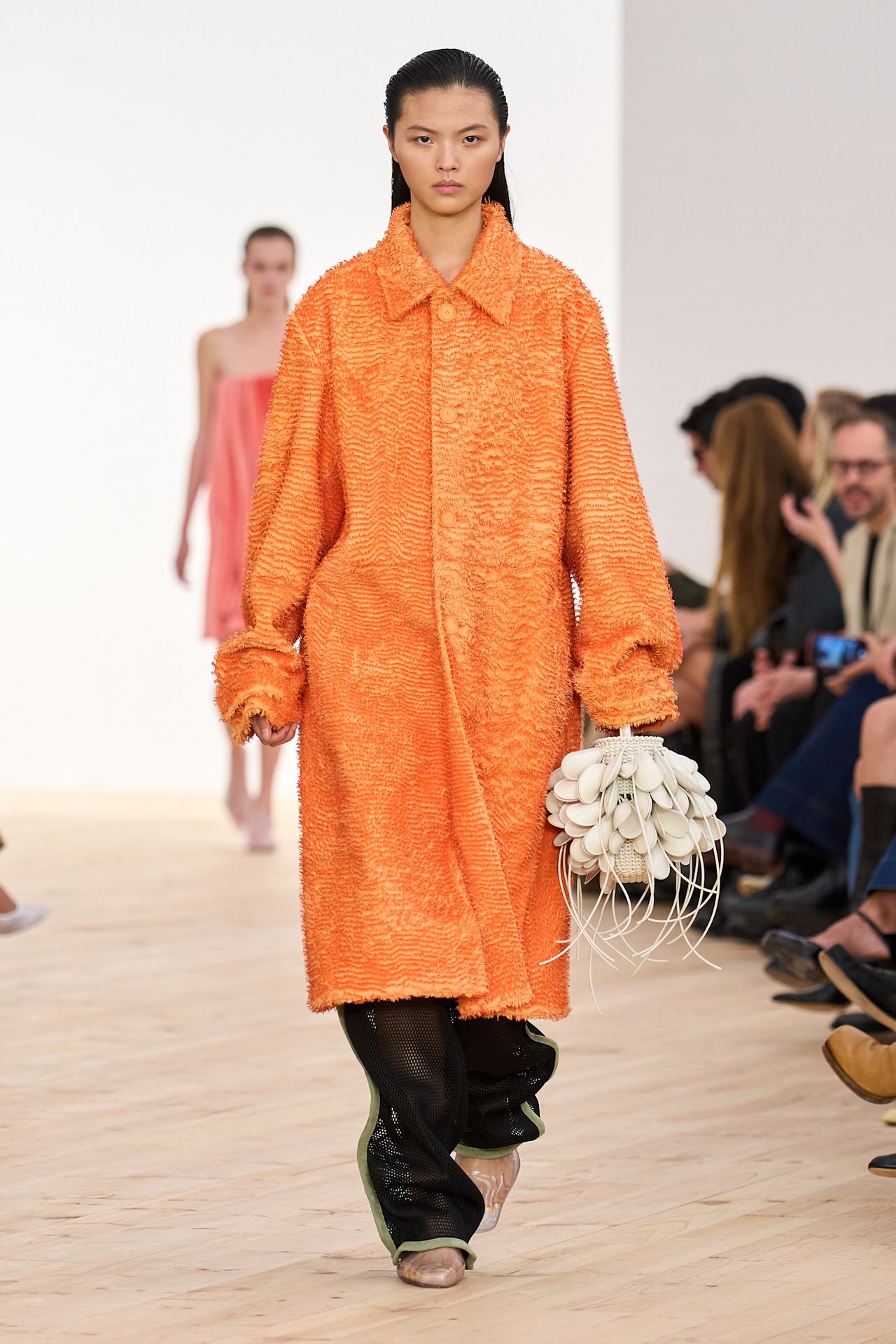 A Loewe model wearing a fuzzy orange coat styled over black perforated leather pants, clear pointed-toe loafers, and a white floral-shaped bucket bag.
