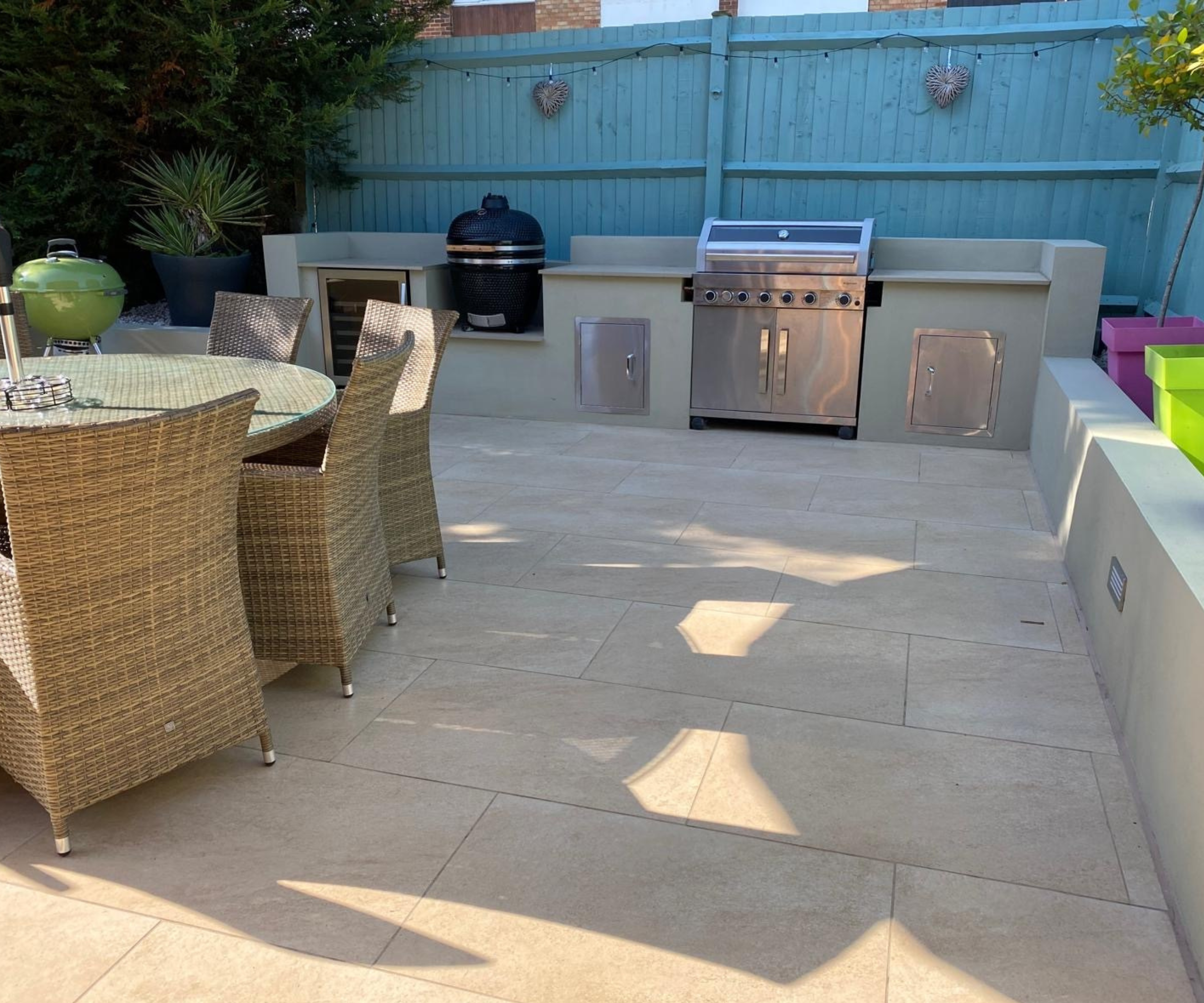 Sam's outdoor kitchen with beige patio slabs, table and chairs to left and worktop to rear, built in kitchen with stainless steel doors and multiple BBQs.