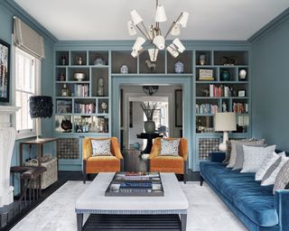 Blue painted large living room space with floor to ceiling storage and display units, two orange fabric chairs and blue sofa with gray cushions, large gray rug an textured gray coffee table and ottoman, fireplace with artwork hanging above, lantern modern chandlier