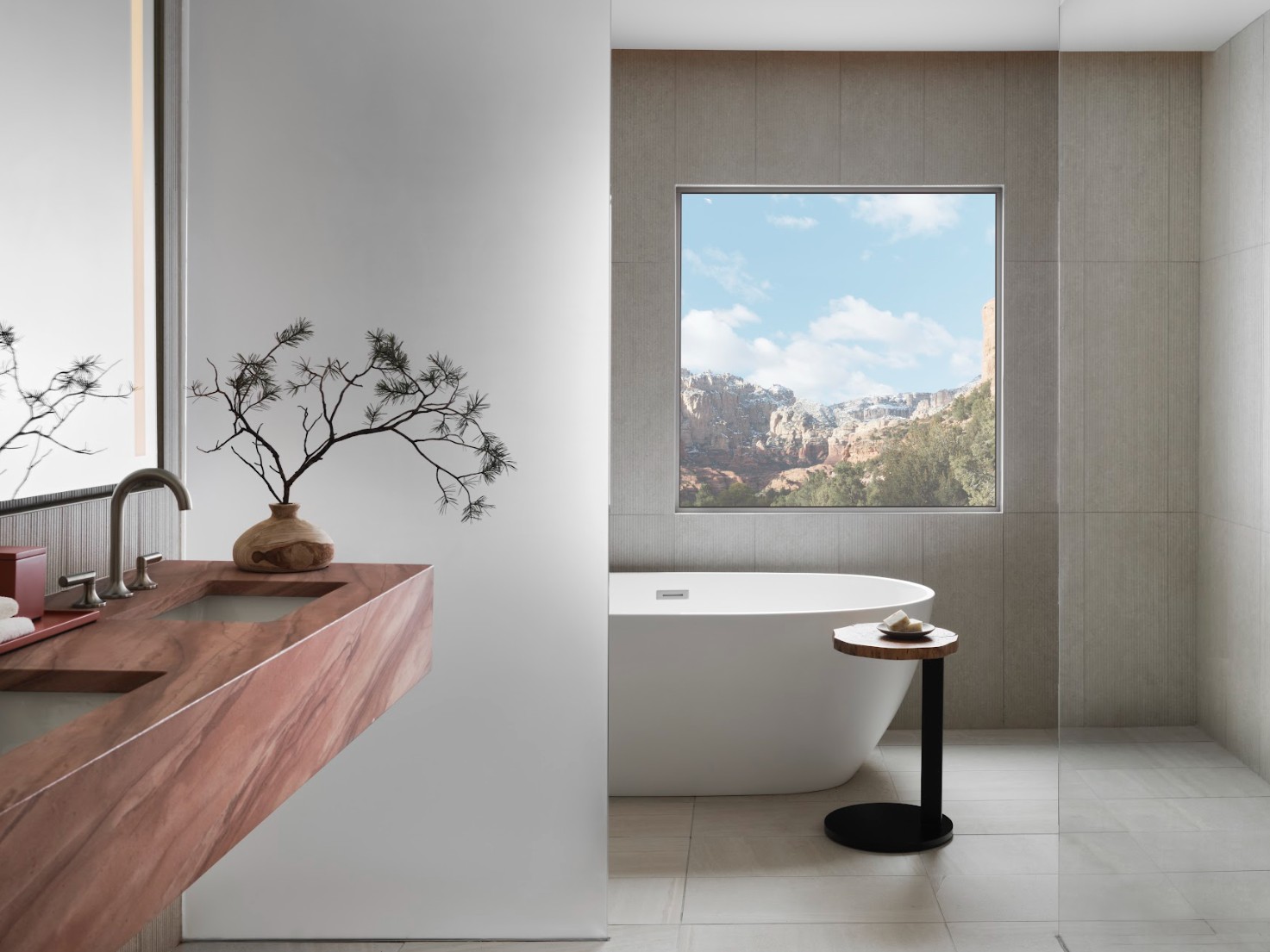 A luxury bathroom at Mii Amo resort and spa. In the foreground is a beautiful, natural-toned bathroom with a bonsai plant on the counter. A large picture window frames a mountain view.