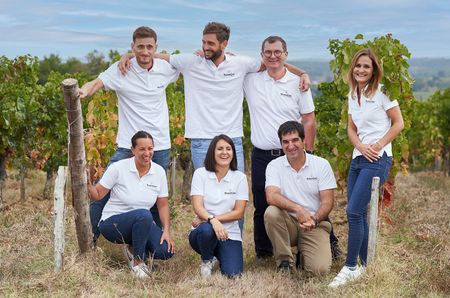 Seven people in white polo tops standing and kneeling in front of green vines