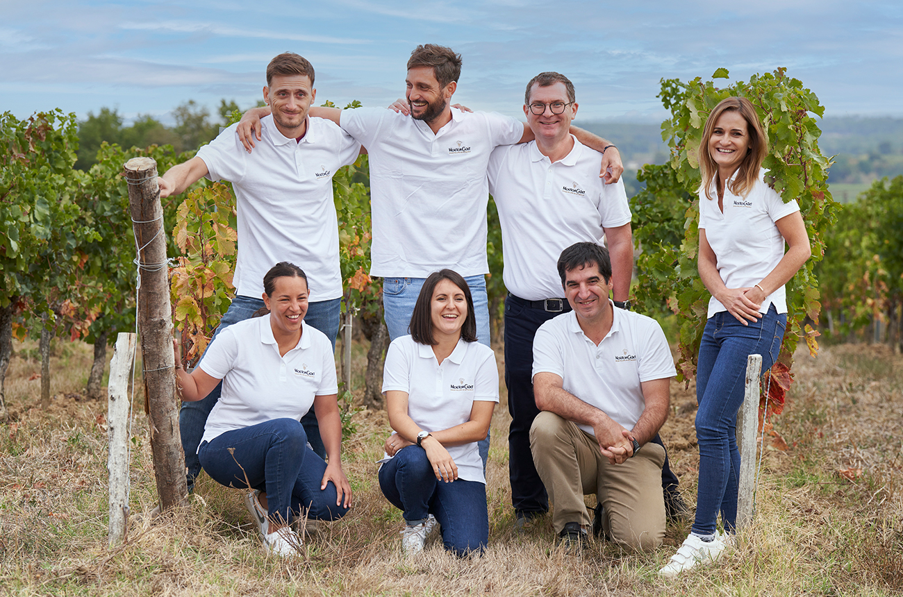 Seven people in white polo tops standing and kneeling in front of green vines