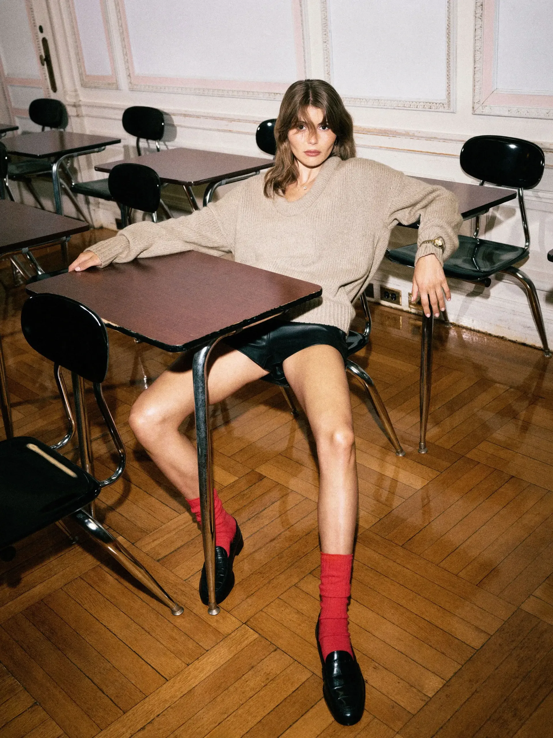 Olivia Jade sits at a school desk wearing the Reformation Jonie jumper, silk shorts, red socks and black loafers