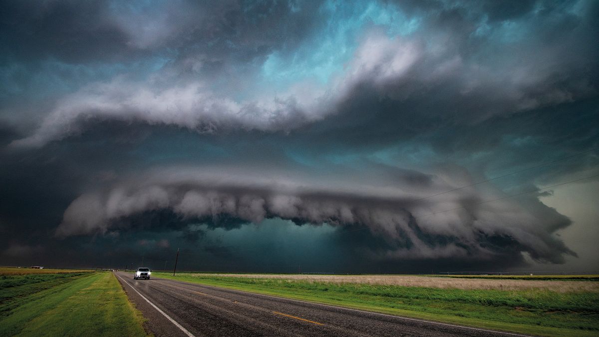 A car driving under a massive dark shelf cloud on the horizon