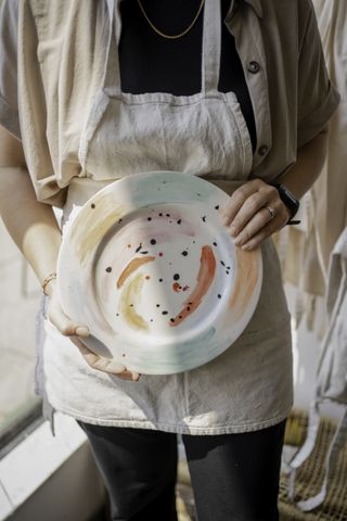 An alumni of Pottery Studio LDN holds the plate she has just decorated, which glows in natural light and bright hues.