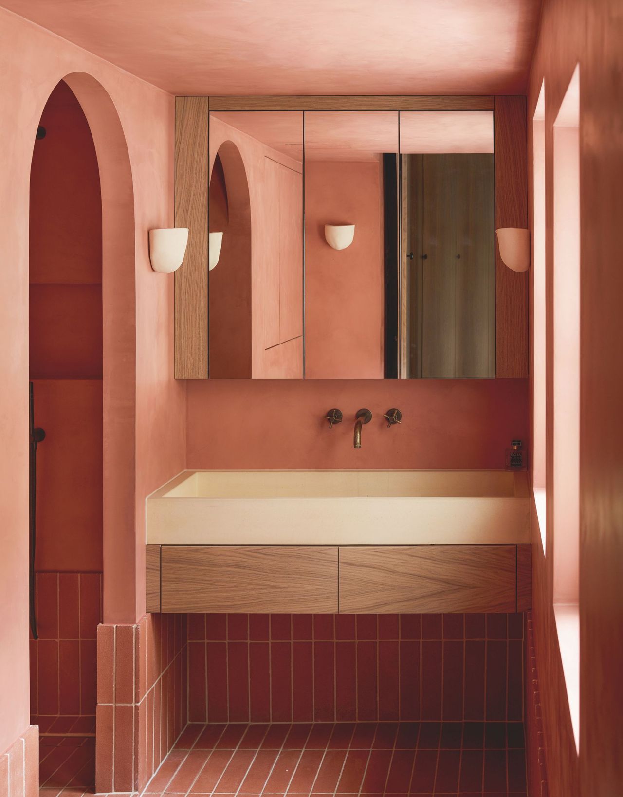 A modern pink bathroom with an arched doorway leading to a shower and a large cream sink with two wooden drawers above pink tiles that run from the floor up to them and a built-in mirror unit