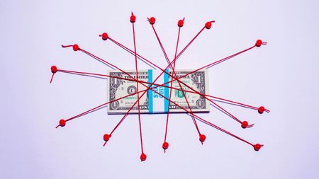 Bundle of US $1 bills tied down on white surface with bright red string and red thumb tacks