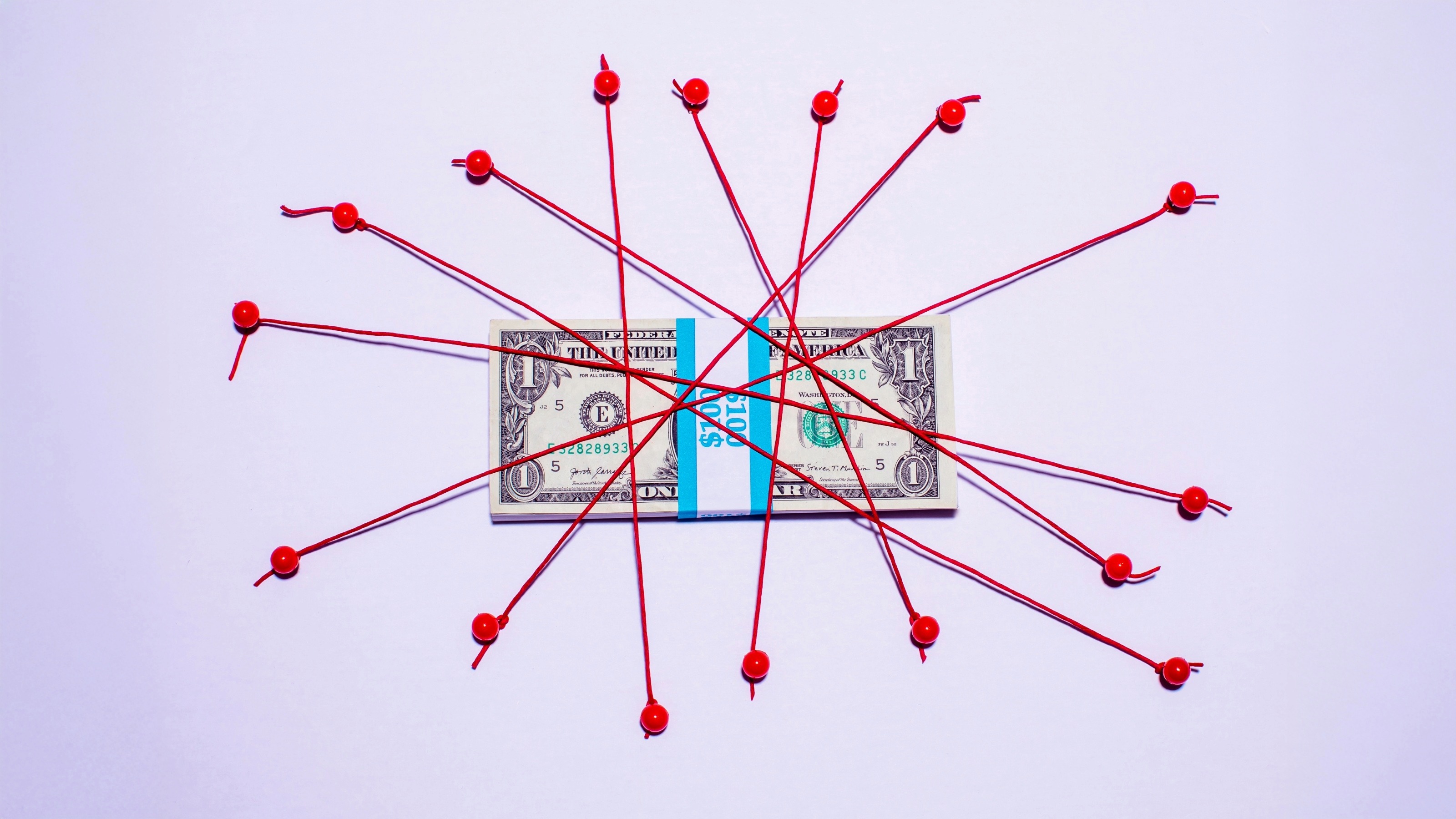 Bundle of US $1 bills tied down on white surface with bright red string and red thumb tacks