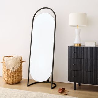 Folded Ellipse Metal Standing Mirror with black frame standing in bedroom beside chest and with a storage basket on the other side