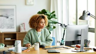 Woman sits at a desk in a brightly lit room, looking at a computer screen.