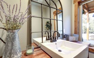 Arched mirro over a square white bathroom sink, with a metal jug of lavender