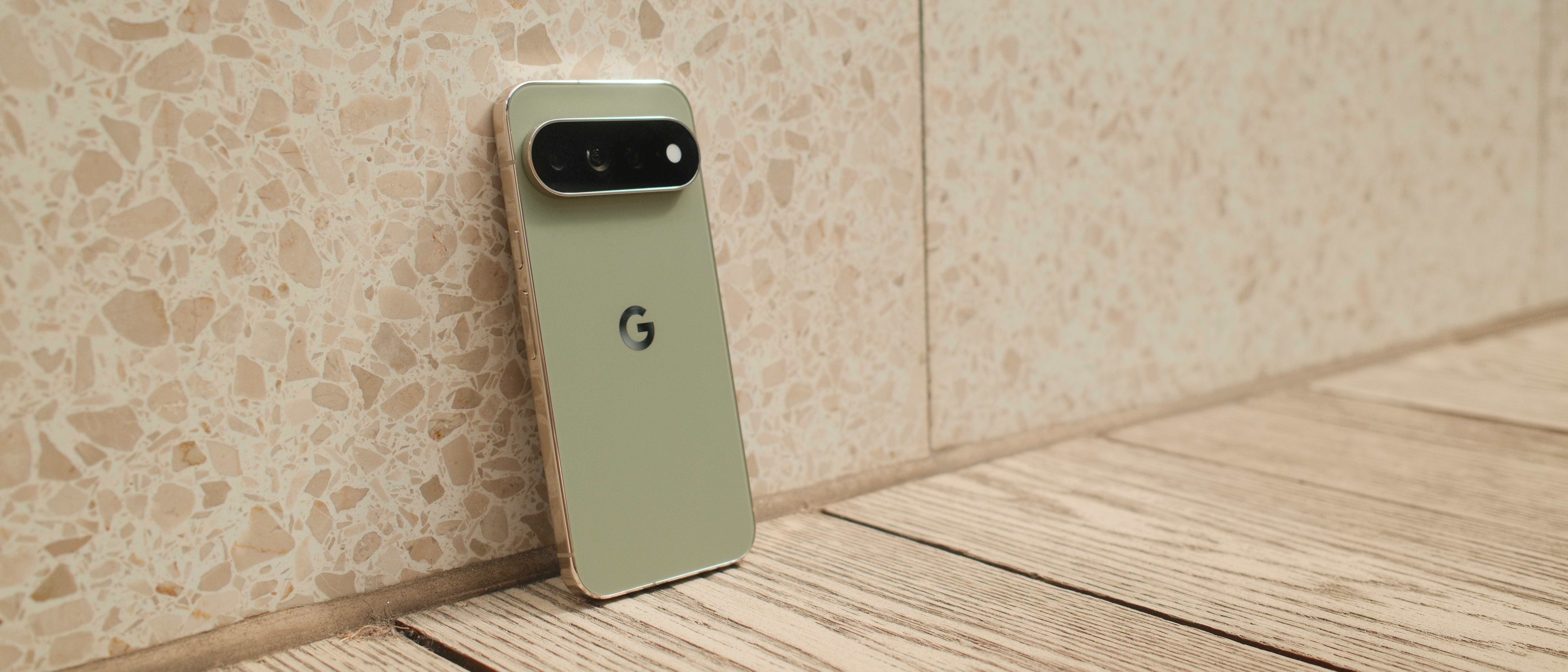 Google Pixel 11 Pro XL leaning against a light stone wall on a wooden floor, showing its rear design and camera bar.