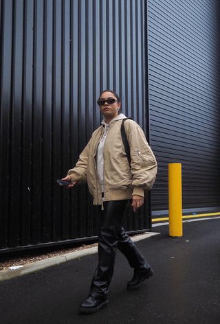 A woman walking outside wearing a beige hoodie under a matching bomber jacket with black leather trousers and black ankle boots.