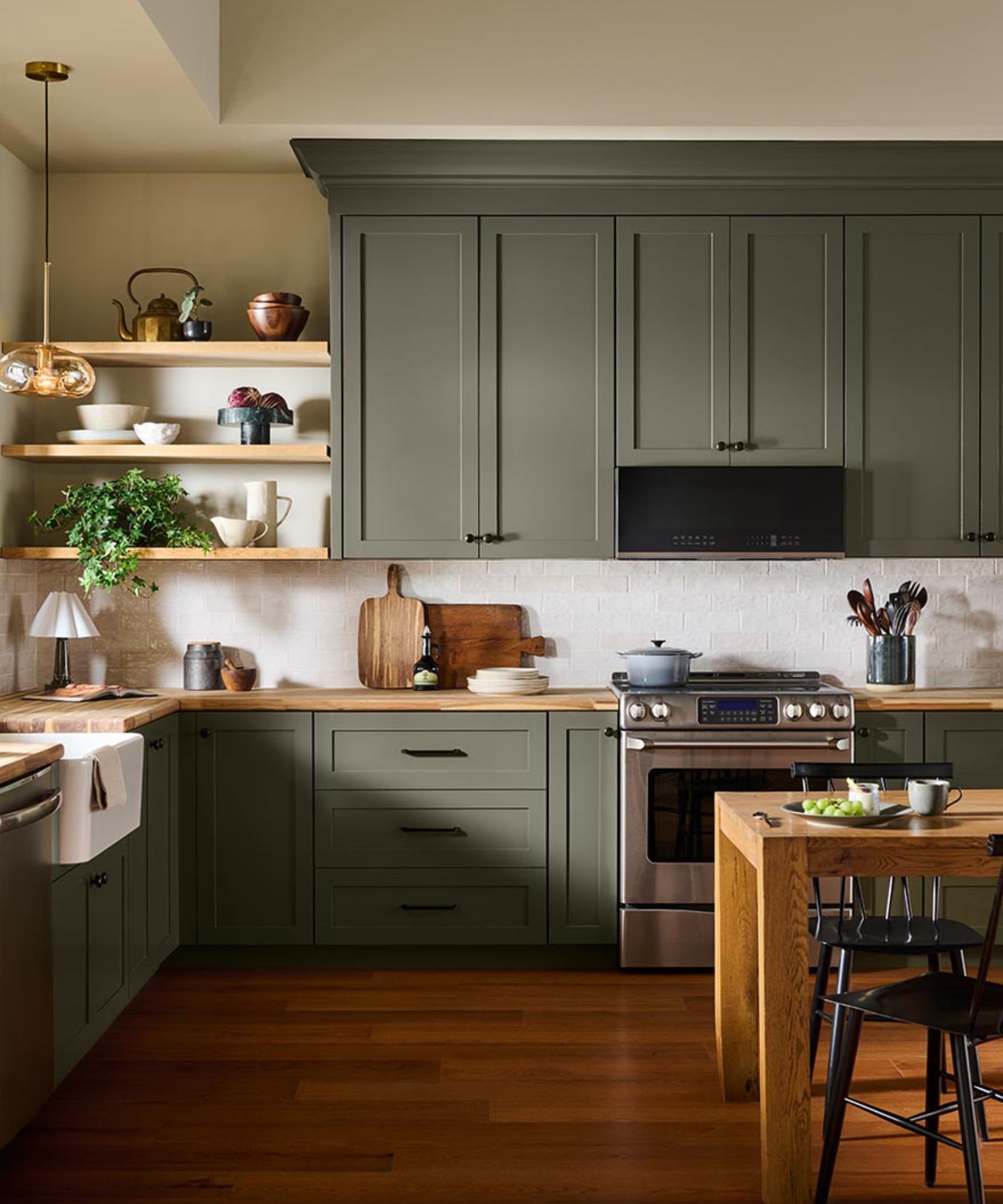 Kitchen with olive green cabinetry, open wooden shelving with ceramics, a wooden island with black spindle chairs and a farmhouse sink.