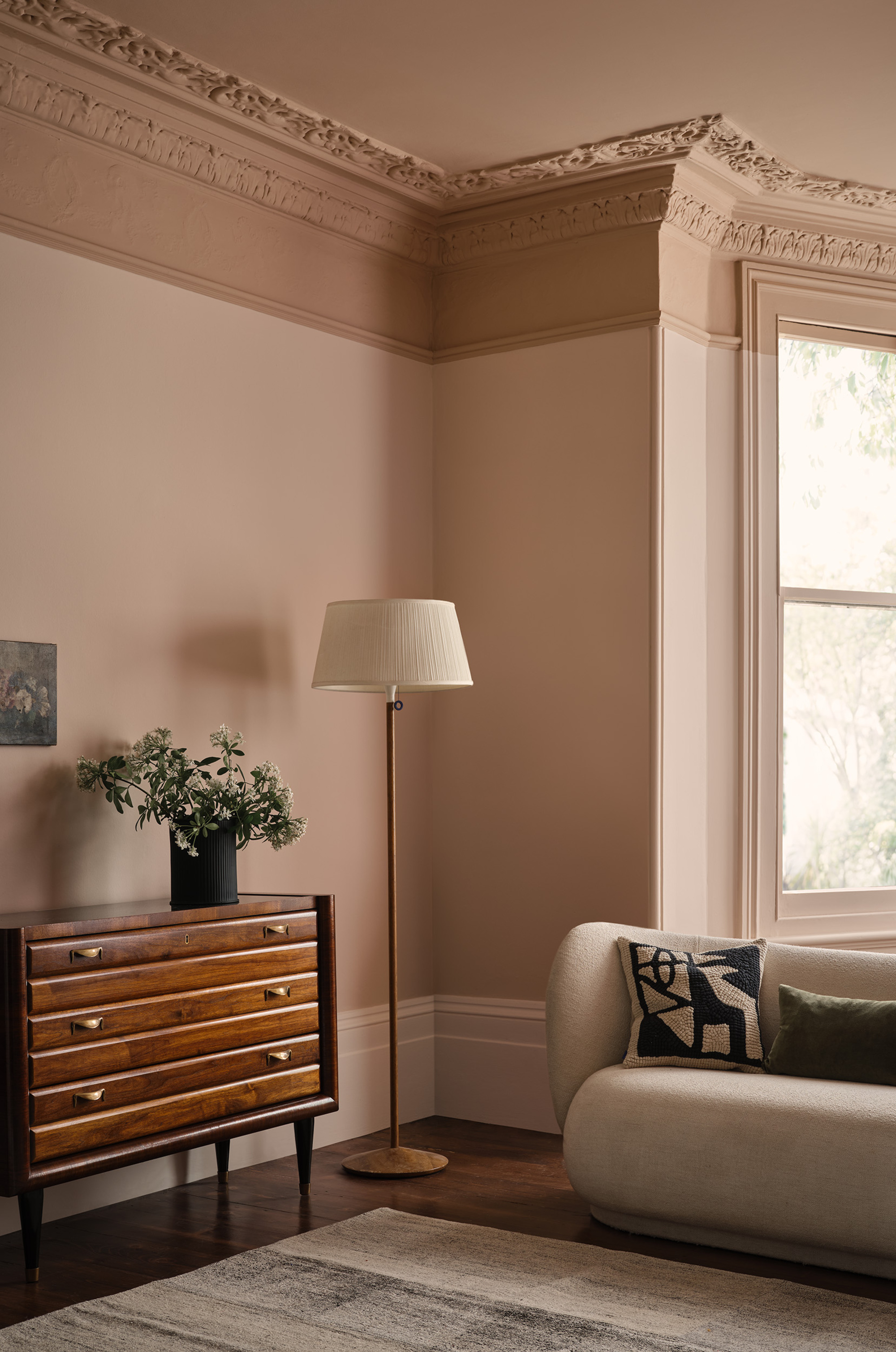 living room with curved sofa styled with cushion, floor lamp next to a console with a vase of flowers, pink walls with a darker shade of pink on the cornice and ceiling