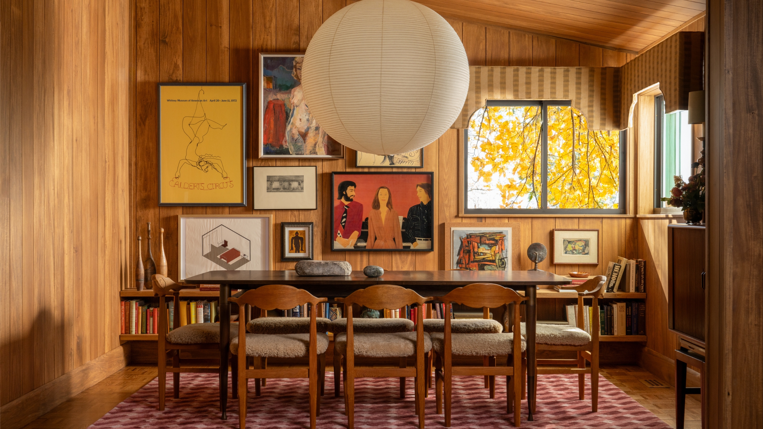 A mid-century dining room with a sloped ceiling paneled in wood to match the walls, and long rectangular dining table, and collected art on the walls