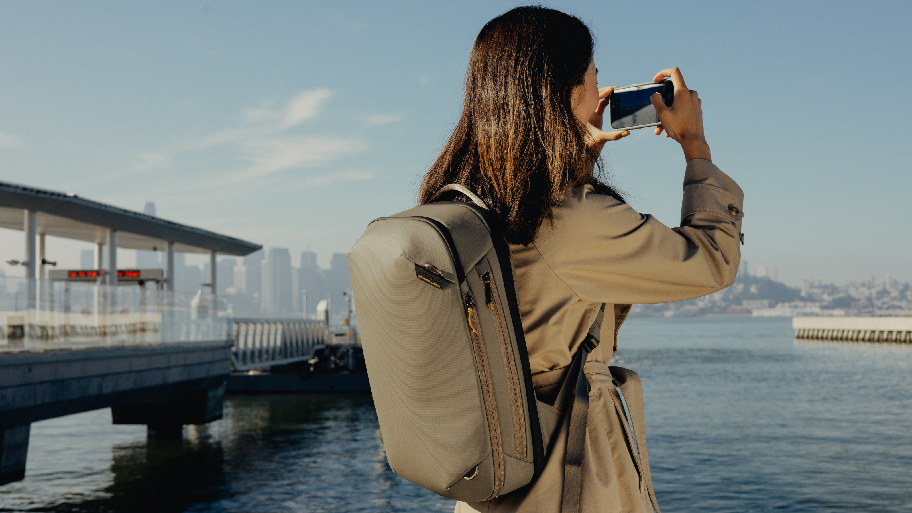 Photograph of a woman wearing the Peak Design Travel Backpack 20L in Stone color, taking a photo of a river harbour with her phone