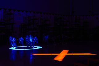 A blue O and a red X are illuminated on the floor of a pitch-black room, as a group of people walk across the O, in 'Squid Game' season 2.