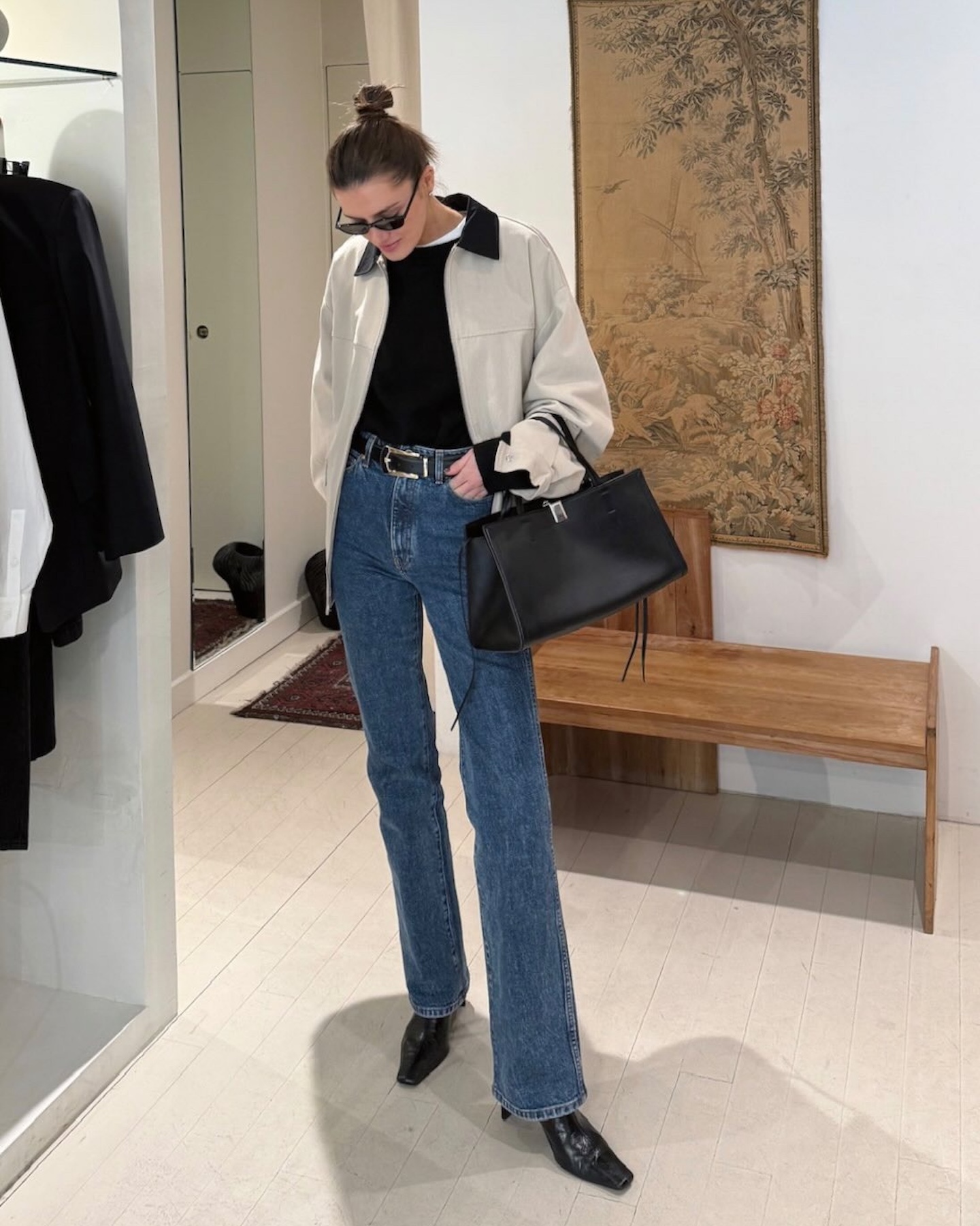 NYC style influencer Coco Schiffer poses in a store wearing a barn coat, simple black bag, black crewneck sweater layered over a white t-shirt, a belt, and jeans