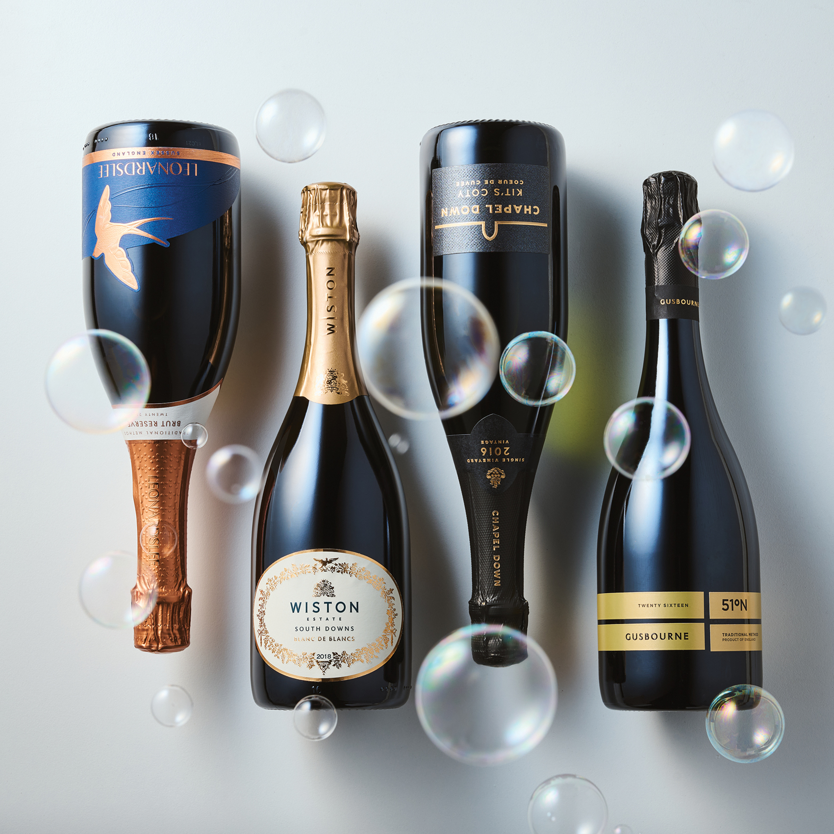 Four bottles of sparkling wine, two upside down, surrounded by floating translucent bubbles on a light background
