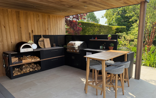 Outdoor kitchen underneath a wooden panelled pergola, featuring black cabinets, log store and a bench for eating