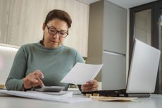 Woman in early 50s looks at finances as she uses calculator at kitchen table.