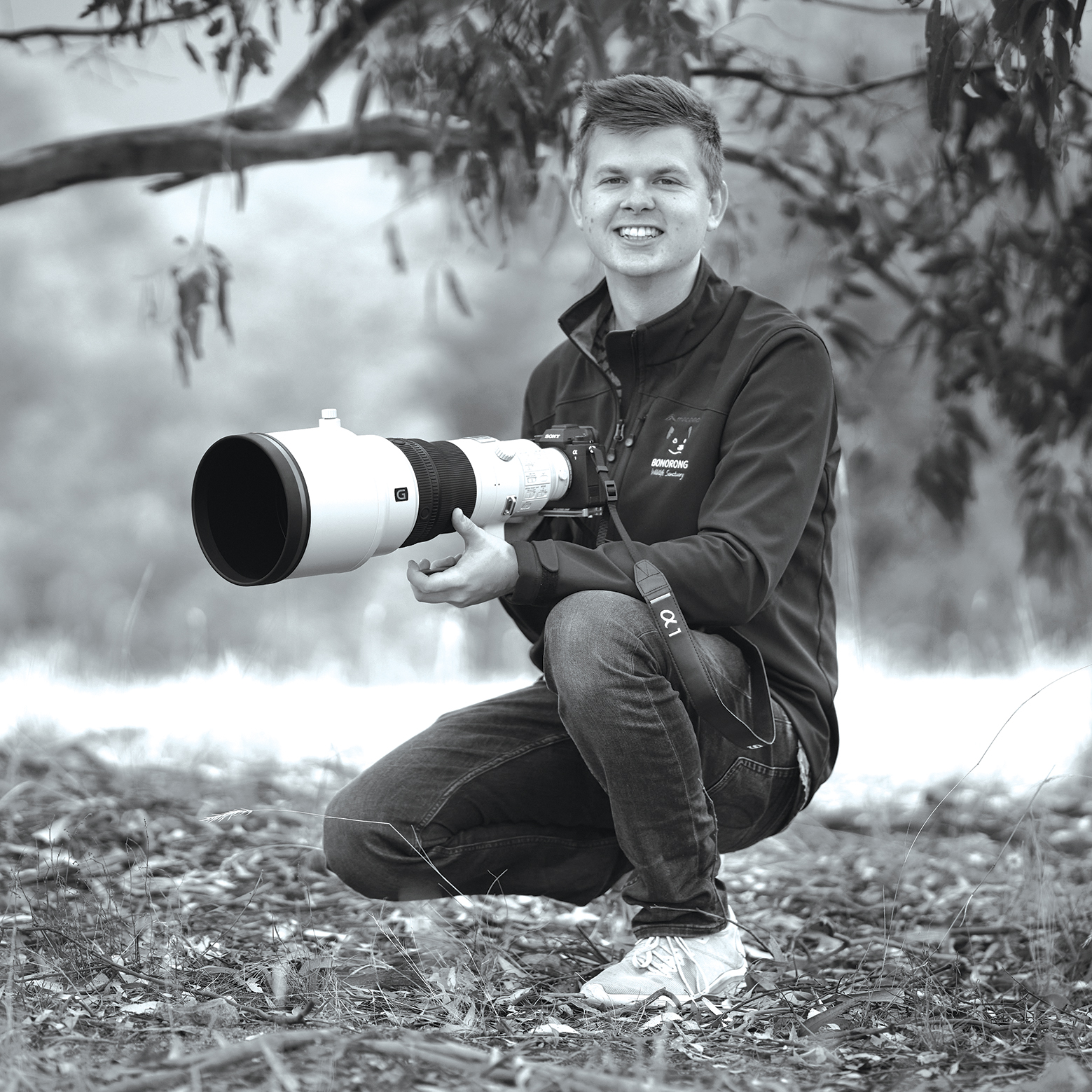 A photographer crouches in nature, holding a large camera with a long lens, dressed in a black jacket and casual jeans