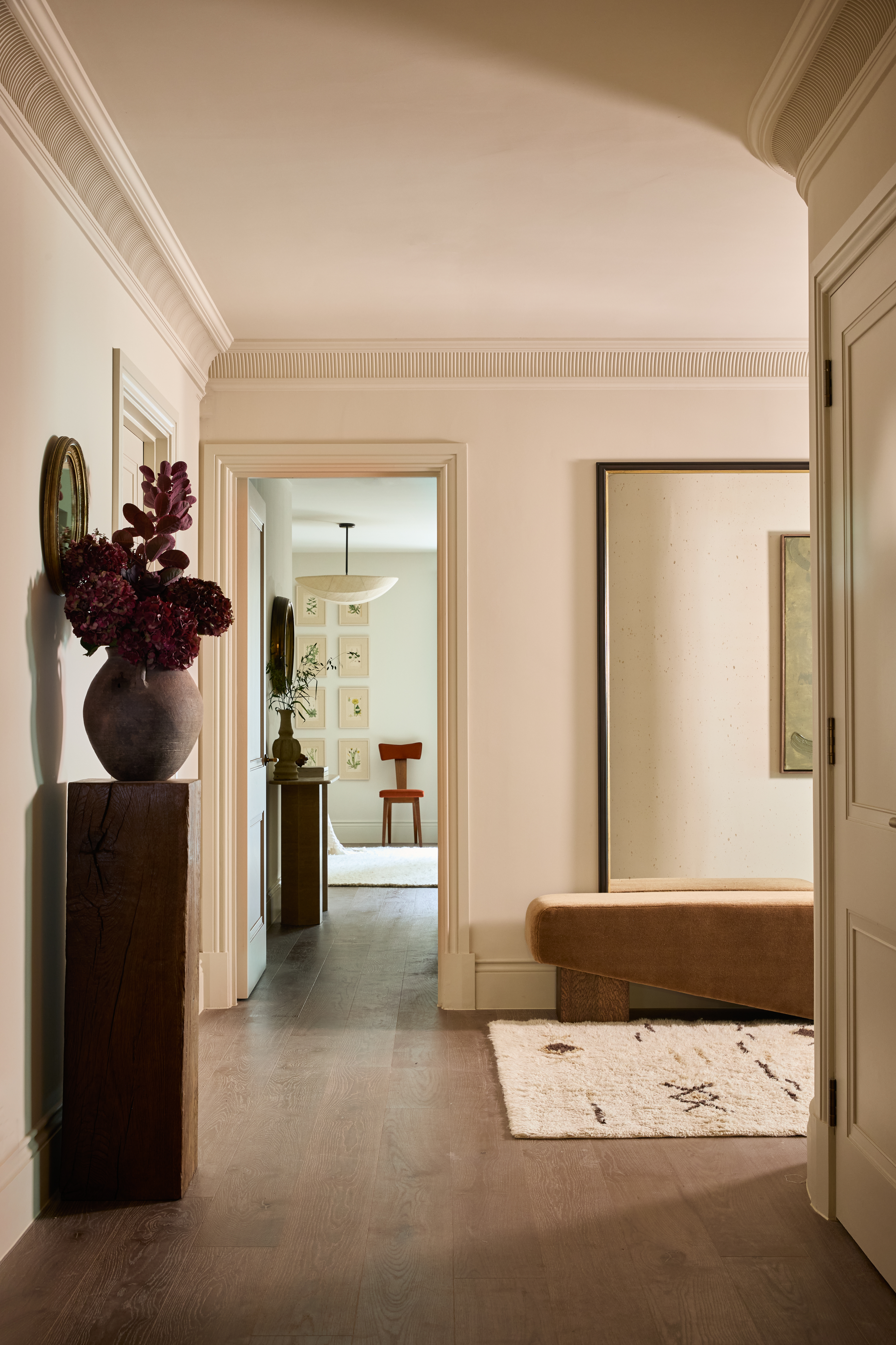 neutral hallway with cream walls, cornicing, timber floorboards, a beige rug, a huge mirror, a velvet bench, a timber plinth with a vase of flowers on top