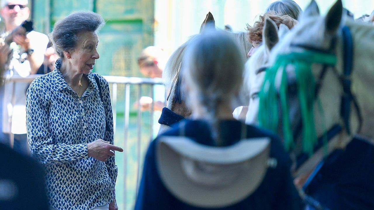 Princess Anne wearing a blue patterned blouse pointing at three horses as women stand in the background with blue shirts