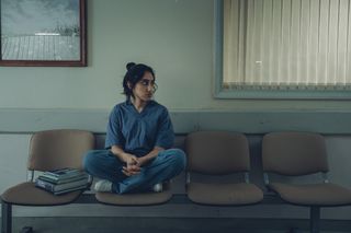 Shruti (Ambika Mod) sits cross-legged on a row of four armless chairs in a hospital corridor, wearing her blue hospital scrubs and with a pile of medical textbooks next to her