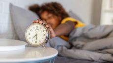 an alarm clock on a bedside table, facing the camera with someone's hand on it. they are lying in bed and reaching out to the alarm clock. 