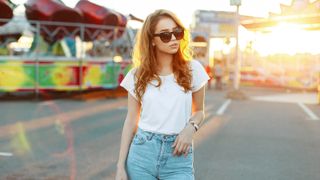 woman in stylish sunglasses in a trendy white t-shirt in blue trendy jeans posing outdoors on the background of carousels