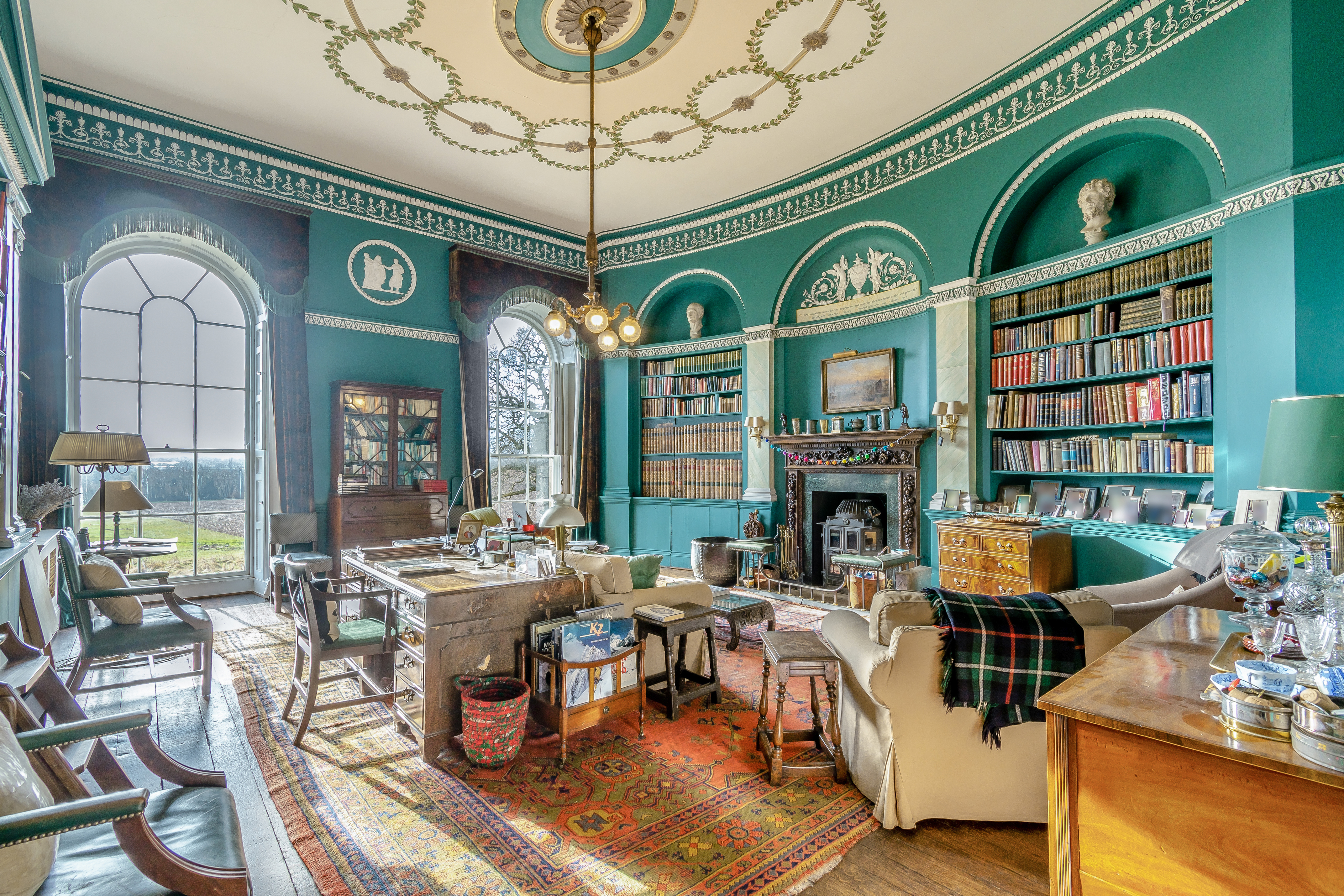 A teal library with an ornate ceiling and many colourful books and pieces of furniture scattered about