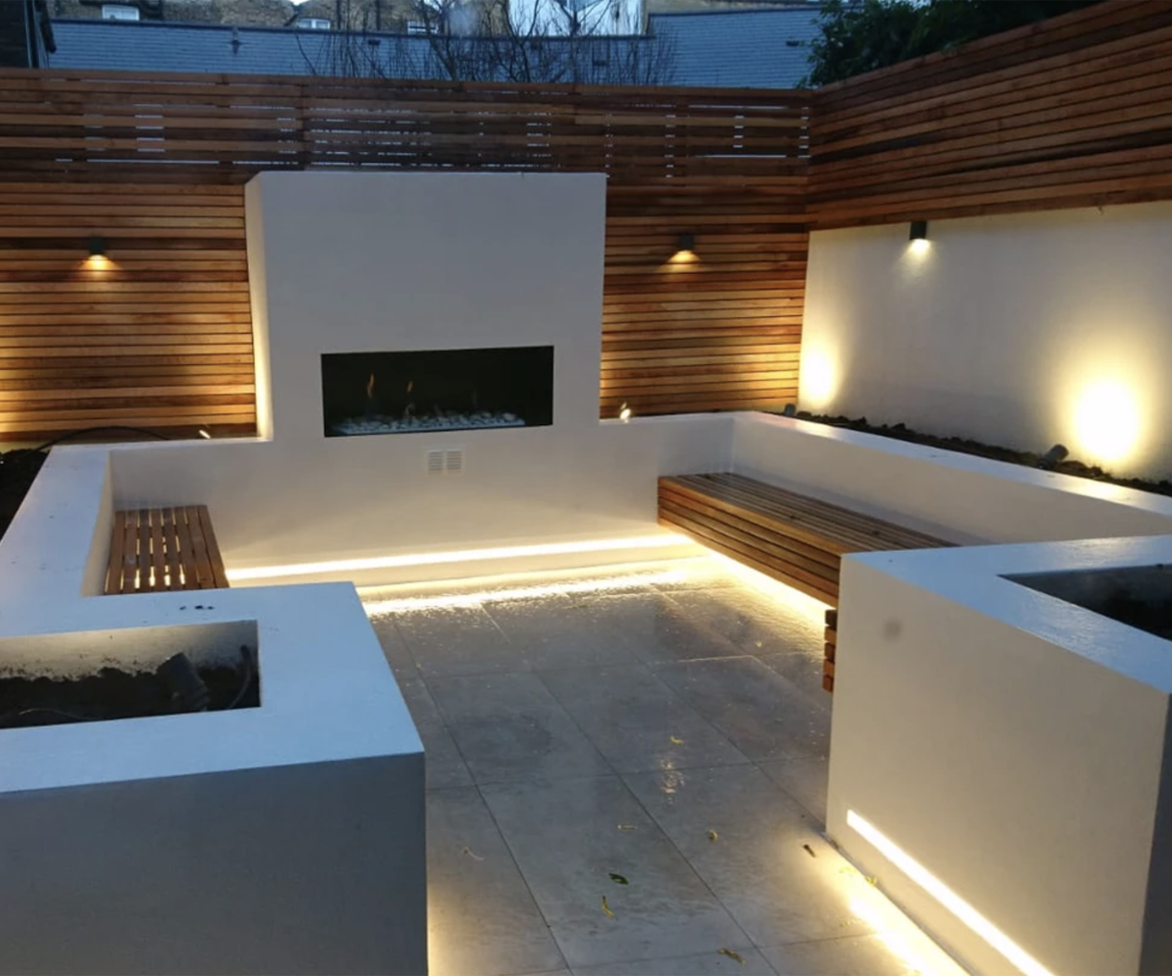 A tiny courtyard garden designed with white builit in seating and raised beds as well as warm uplighting and an outdoor fireplace