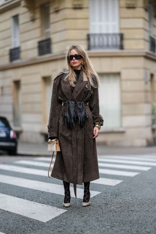 Virginie Conte wears black sunglasses, a dark brown top, a brown suede trench coat, a brown suede skirt, a black leather belt, black leather gloves, gold bracelets, a white leather Kelly Hermes bag, black lace tights with floral details, brown leather Gucci shoes high heels, during a street style fashion photo session, on November 30, 2024 in Paris, France