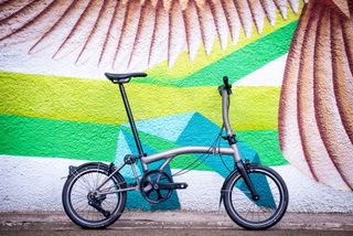 A silver Brompton T-Line Urban titanium folding bike leans against a multi-coloured backdrop