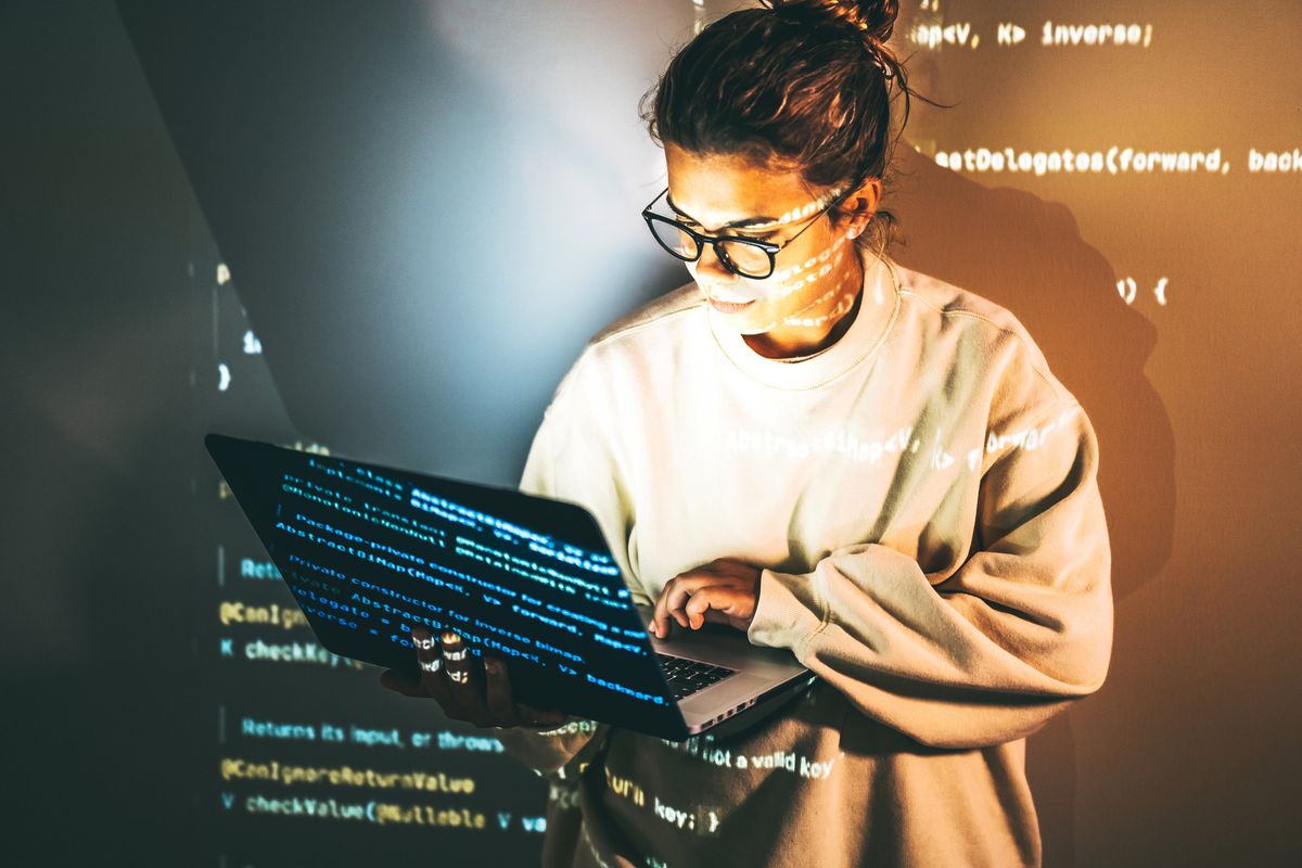 A woman stood typing on a laptop while in front of a projector casting an image of computer code