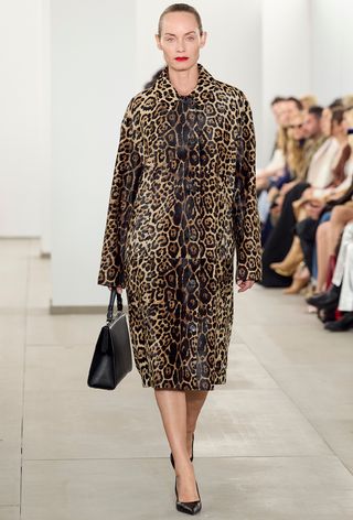 Leopard print coat trend is shown on the fall runway 2024 collection of Michael Kors with a model walking wearing a leopard pony hair coat with black pumps and a black frame bag