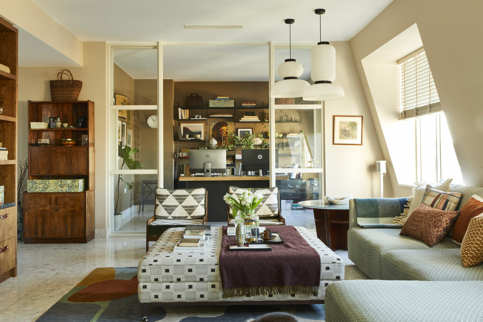 A stylish, modern living room with a cozy sofa, patterned ottoman, two geometric armchairs, glass partition wall leading to a home office, and warm neutral decor with paper pendant lights