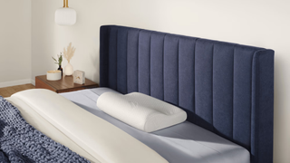 A curved white pillow placed in the center of a blue with blue and white bedding and a side table next to it 