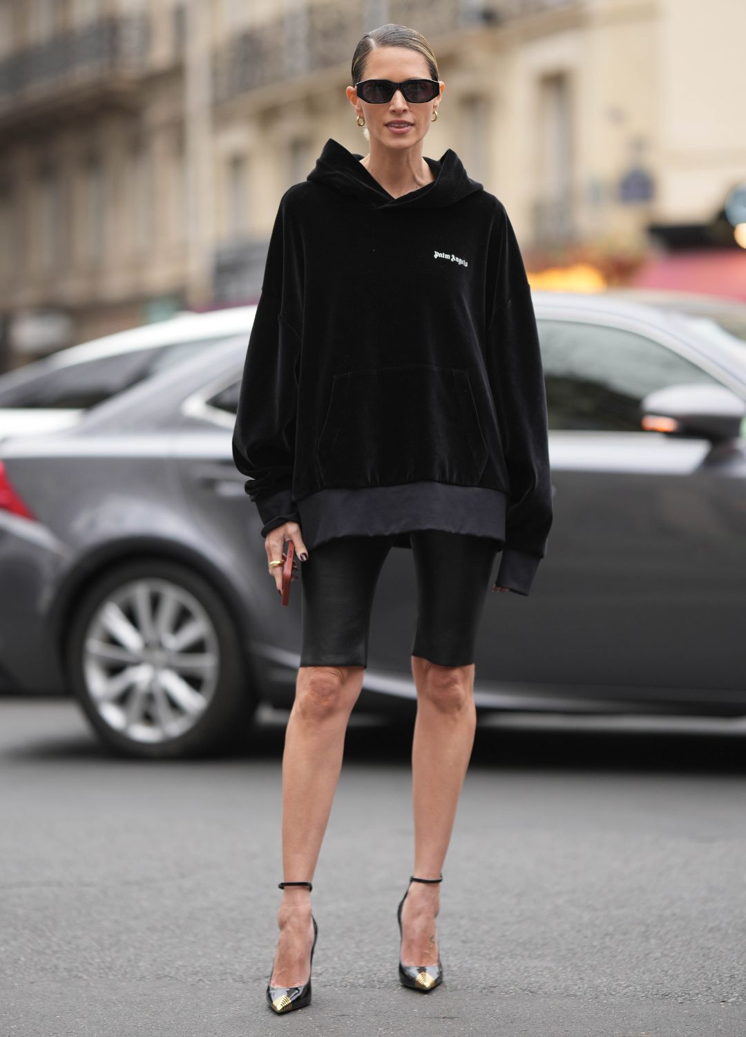 Helena Bordon wears black sunglasses, gold earrings, a black hoodie sweater from Palm Angels, black shiny leather cyclist shorts, black shiny varnished leather with gold metallic toe-cap / pointed pumps heels shoes , outside Palm Angels, during Paris Fashion Week - Womenswear Fall Winter 2023 2024, on March 05, 2023 in Paris, France