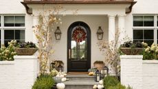 white house with large porch with pillars, a black glass arched door with two wall lights decorated for fall with wreaths, pumpkins and fall foliage