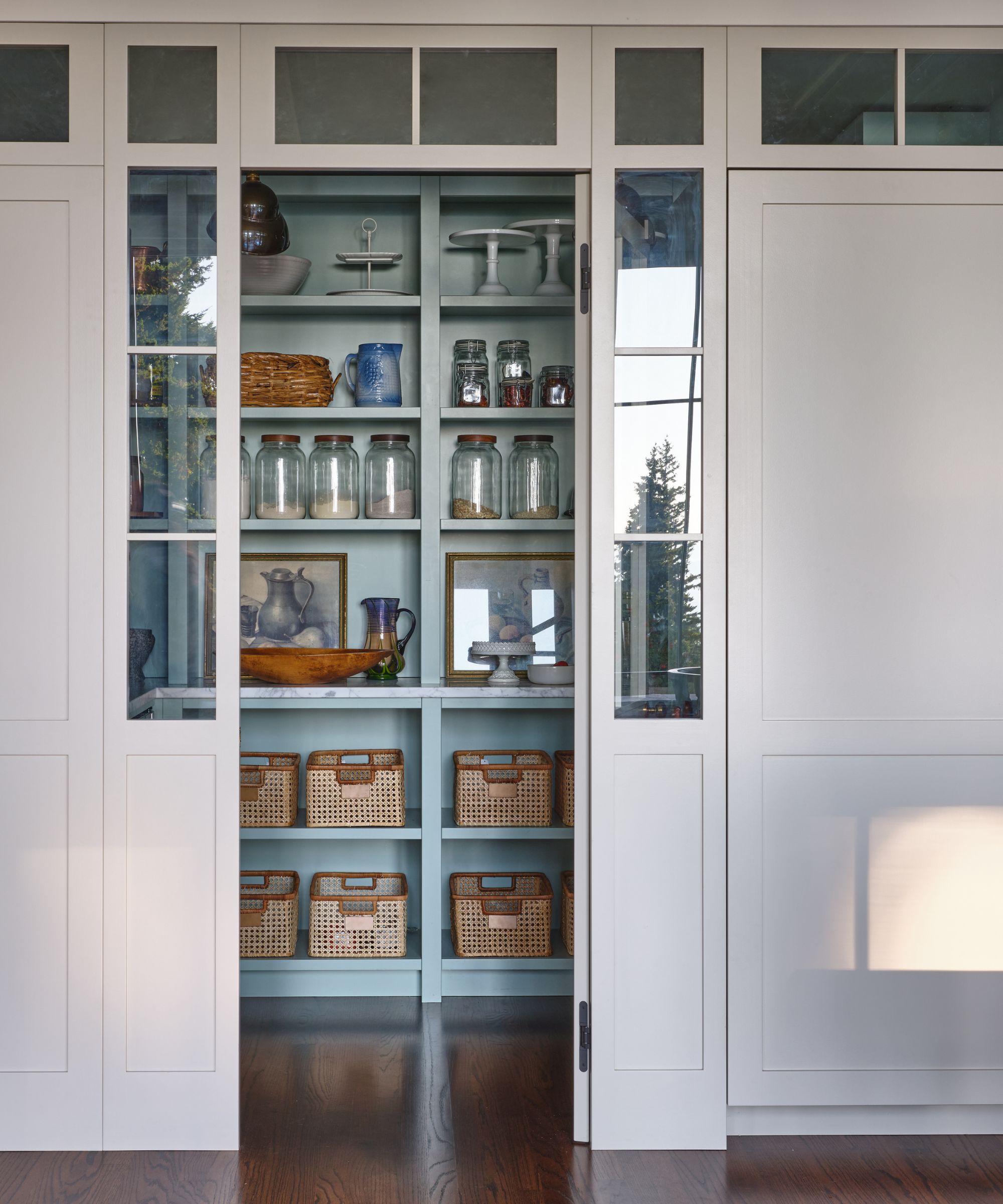 A built-in pantry seen through white glass-paneled sliding doors, featuring pale blue shelves stocked with glass jars and woven storage baskets