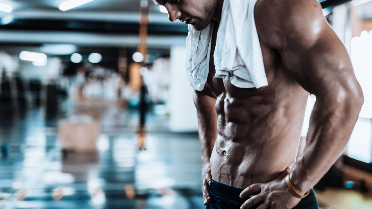 Man standing in a gym topless with a towel round his neck