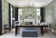 a green wallpapered living room by Marie Flanigan with large windows, a fireplace, neutral furniture and a dark wooden console table at the back of the couch with a navy linen ottoman beneath