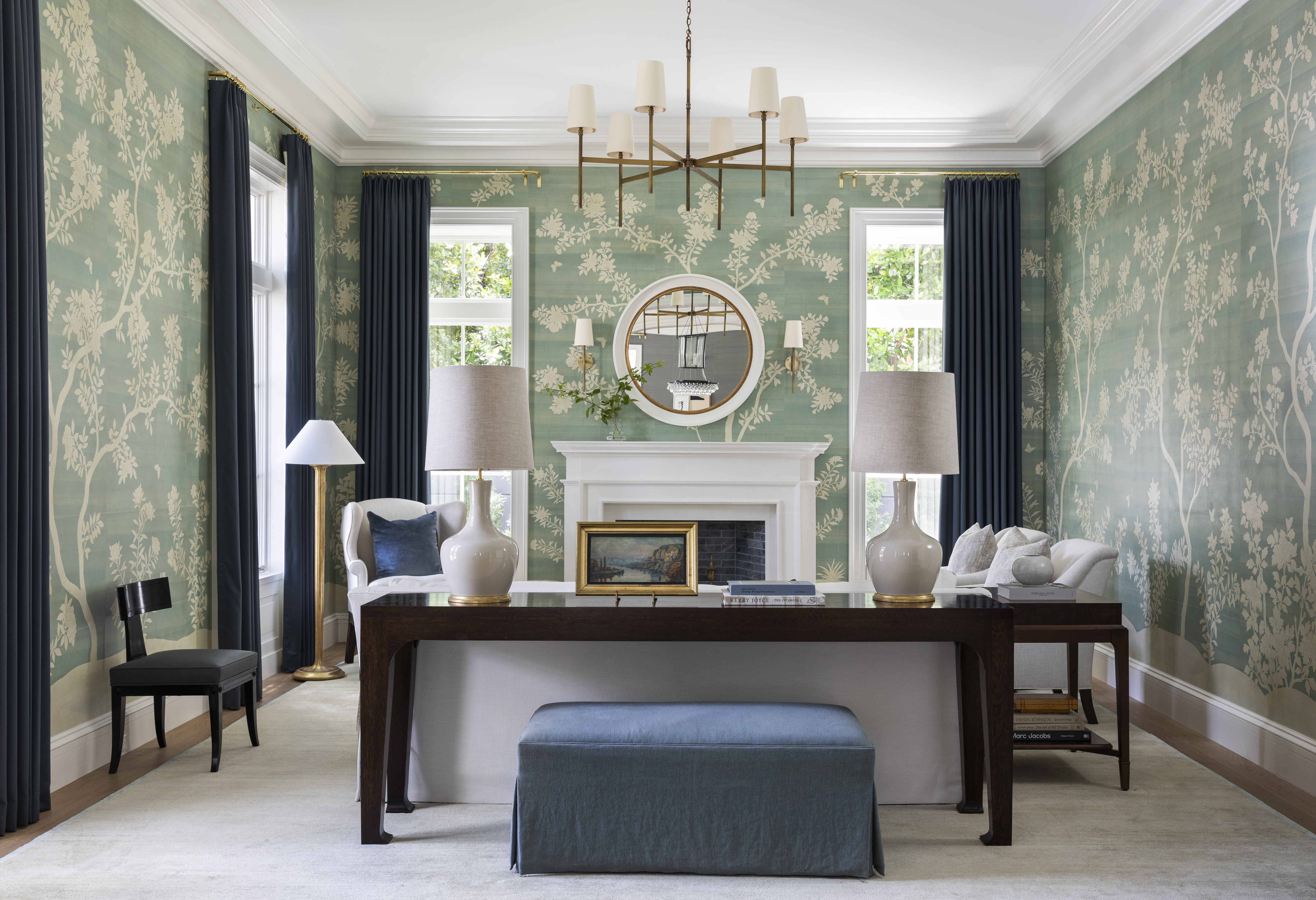 a green wallpapered living room by Marie Flanigan with large windows, a fireplace, neutral furniture and a dark wooden console table at the back of the couch with a navy linen ottoman beneath